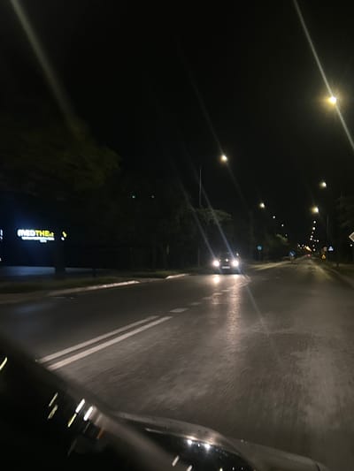The image depicts a nighttime scene on a wet road, likely in Siedlce, Poland, given the reference. A car with illuminated headlights is visible in the distance, driving on the right side of the road. Streetlights line the road, casting pools of light on the wet asphalt. Trees are present on the left side of the road, partially obscuring a lit sign that reads "MEDTHE&." The perspective suggests the image was taken from inside another vehicle. The overall impression is that of a slightly blurred, atmospheric night drive on a suburban or city street.