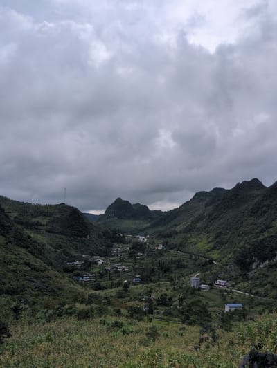 The image depicts a valley in Sà Phìn Commune, Vietnam, under a cloudy sky. The landscape is dominated by lush green, tree-covered mountains that frame a village nestled within the valley. A winding road is visible, connecting various buildings and houses scattered throughout the settlement. In the foreground, tall, dry grass and vegetation dominate the scene. On one of the distant hilltops, a tall, slender tower stands prominently. The architecture of the buildings suggests a rural setting, with some structures appearing to be multi-story. The overall impression is of a remote and verdant mountain community.