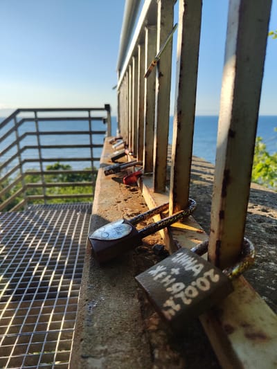 The image depicts a view from an elevated platform overlooking the Baltic Sea in Valaste, Estonia. In the foreground, a metal railing with visible signs of rust runs along a concrete ledge. Several padlocks are attached to the railing and the ledge, with some showing inscriptions. A metal grated walkway is visible to the left of the image. The background features the blue expanse of the sea under a clear sky, with green foliage visible on the right side. The lighting suggests it is either early morning or late afternoon, with long shadows cast by the railing. There are no people or animals present in the frame.