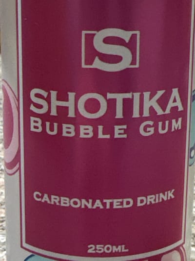 The image displays a close-up, vertical shot of a magenta-colored beverage label for a "SHOTIKA BUBBLE GUM Carbonated Drink," situated in Dabouq, Jordan. The label features a white stylized 'S' logo at the top within a square bracket design. Below this, "SHOTIKA" is printed in large white capital letters, followed by "BUBBLE GUM" in smaller white capitals. Further down, "CARBONATED DRINK" is visible in white capital letters, presented on a slight upward curve. The volume "250ML" is indicated at the bottom. The left periphery of the label shows a partial white border with a pink concentric curve pattern, while the right edge displays a plain white border. The background behind the container is an indistinct, light-colored, grainy, and out-of-focus textured surface. No individuals or specific actions are discernible.