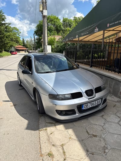 A silver car with license plate "OB 7865 BX" is parked on a street in Bratya Daskalovi, Bulgaria. The car is parked next to a sidewalk and a building with a black metal fence and a green awning. Trees and utility poles line the other side of the street. The sky is visible between the trees and shows some clouds.
