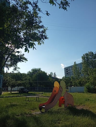 The image depicts a sunny day in a playground setting. A colorful, elephant-shaped slide with orange and yellow elements is the central object. The slide appears to be made of wood and plastic and is situated on a grassy area. In the background, a residential building with multiple stories and balconies stands tall, surrounded by lush green trees. A dark-colored car is parked near the trees to the left of the playground. A bench is visible on the grass, and a portion of a fence runs along the middle ground. The sky is clear and bright blue, suggesting it is daytime, likely late morning or early afternoon, given the strong sunlight casting shadows. The overall atmosphere is peaceful and reminiscent of a typical urban residential area in Moldova, specifically Rezina, during warm weather. There are no people actively playing on the slide or in the immediate vicinity, contributing to a tranquil scene. The lush green grass and vibrant foliage indicate a pleasant season, likely spring or summer.