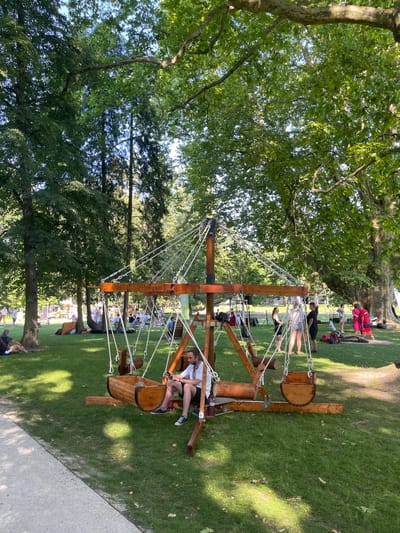 A man sits on a wooden swing designed to resemble a boat in a park on a sunny day. The swing set is made of wood and rope and has a circular design with multiple swings. In the background, there are trees, people sitting on the grass, and a few tents or stalls, suggesting an outdoor event or festival. The location is identified as the city of Stupava, Slovakia.