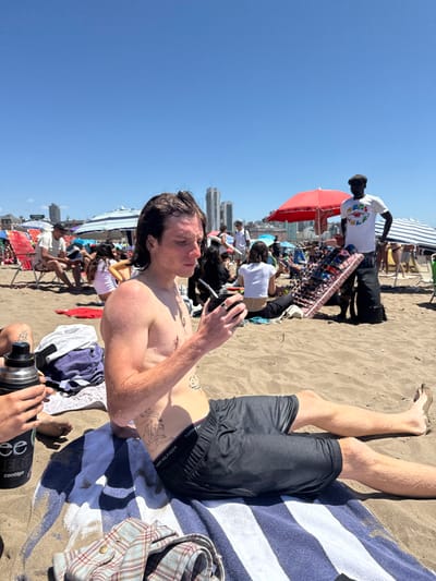 A man in black shorts lies on a striped towel at a crowded beach. He is shirtless and holds a phone. A tattoo is visible on his torso. Other people are relaxing on the beach, some under umbrellas. A man in a white shirt stands with a rack of merchandise. Buildings can be seen in the background. A water bottle is visible in the foreground. The scene is sunlit.