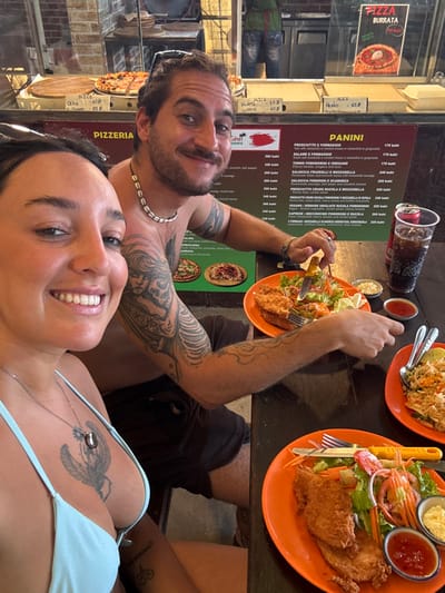 A couple takes a selfie at a pizzeria in Thailand. They are seated at a dark wooden table with plates of fried chicken salad, rice, and condiments. A tall glass of cola is also visible. The man is shirtless with tattoos and a shell necklace. The woman wears a light blue bikini top and a necklace. Behind them is a menu board listing pizza and panini options in Thai baht, along with various pizzas on display.