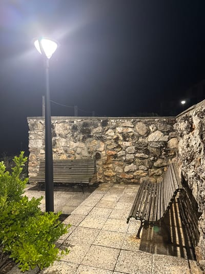The image depicts a nighttime scene in Tyulenovo, Bulgaria, featuring a stone wall and paved area illuminated by a street lamp. In the foreground, to the left, is a dense green shrub. A black lamppost stands near the shrub, casting bright light. Two wooden park benches are present. One bench is positioned in front of the stone wall, to the right of the lamppost, and the other is partially visible to the right of the frame. The stone wall is constructed of irregular, roughly hewn stones of varying sizes and colors, with some patches of dark discoloration. The ground is paved with square tiles, and a shadow from the lamppost and the rightmost bench falls across it. The sky above is dark, indicating nighttime. There are no people or animals visible in the scene.