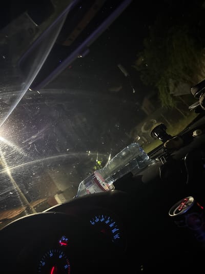 This media file shows the interior of a car at night. The windshield is visible, reflecting trees and streetlights from outside. On the dashboard, a speedometer and a temperature gauge are illuminated, with the speedometer showing readings up to 220 and the temperature gauge showing a normal operating temperature. A partially filled plastic water bottle is attached to the windshield with a suction cup mount, likely holding a dashcam or phone. The scene is dark, suggesting it is late at night or early morning. The car appears to be parked or slowly moving, as indicated by the visible dashboard instruments. The presence of a car interior, dashboard, and windshield firmly places the setting within a vehicle. The reflection of the outside world on the windshield adds depth and context, hinting at a drive through a populated area with vegetation. There is no clear indication of any people in the frame. The overall mood is quiet and still, characteristic of a nighttime drive. The visible text includes numbers on the car's dashboard instruments, such as "20", "40", "60", "80", "100", "120", "140", "160", "180", "200", "220" on the speedometer, and "0", "30", "60", "90", "130" on the temperature gauge, as well as "11900".