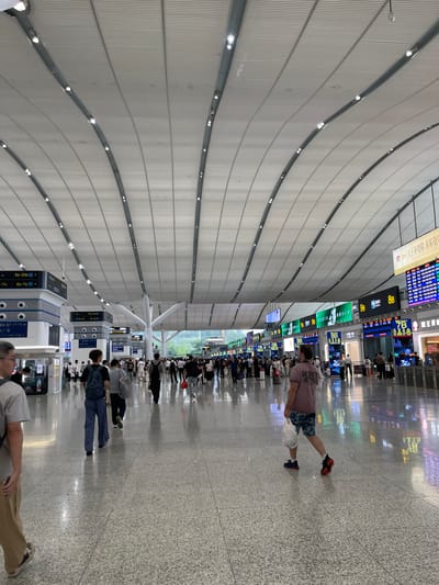 This is a vertically oriented, full-shot photograph taken inside a modern airport terminal. The vast interior is characterized by a curved, white ceiling with numerous lights and parallel ribbed panels, creating a sense of spaciousness. The floor is a polished, speckled terrazzo.
The terminal is populated with numerous people, some walking, others standing in lines, and some seated. Most are casually dressed, suggesting travel. In the foreground, a man is walking away from the camera, wearing a pink t-shirt and patterned shorts, carrying a white bag. Further into the terminal, many people are queuing near information boards displaying flight information in a mix of Chinese and English characters, as well as numbers like "8B", "9B", "7A", "6B-7B", "8A-9A", and "18-20B". Digital screens displaying colorful graphics are also visible. The lighting suggests it is daytime, with bright, artificial illumination throughout the terminal. The atmosphere appears orderly and functional, typical of an airport.