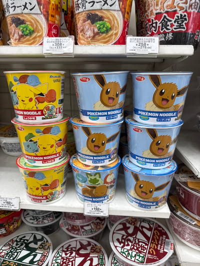 This media file displays a close-up, vertical shot of several shelves stocked with instant ramen cups. The products are primarily themed around popular Japanese anime characters, specifically Pokémon.
The central focus is on rows of "Pokémon Noodle" cups. One stack features Eevee on a light blue background, advertising "Seafood Flavor" and "Nutritional Food (Calcium)." Another stack prominently displays Pikachu and other Pokémon characters on a bright yellow background, advertising "Soy Sauce Flavor" and including a note about a sticker inside. Several of these Pokémon noodle cups have price tags attached, indicating prices such as ¥160 (¥172.80 including tax).
Interspersed with the Pokémon noodles are other brands and flavors of instant ramen. One noticeable cup is branded as "Sakauchi Shokudo Kitakata Honten Kanshou: Kogashi Negi Yuyu Chuka Soba," a "Charred Green Onion Oil Chinese Soba" endorsed by Sakauchi Shokudo, priced at ¥258 (¥278.64 including tax). Another is a "Takuyuu Untenshu Umaiten: Hakuryuuken Kanshou: Tonkotsu Ramen," a "Driver's Favorite: Hakuryuuken Endorsed: Pork Bone Ramen," priced at ¥308 (¥332.64 including tax). In the background, other ramen cups with red and white branding are visible, with some text suggesting a "50th Anniversary" and "Classic" recipe.
The overall scene is likely a retail environment, such as a supermarket or convenience store aisle in Adachi, Japan, given the context. The lighting suggests an indoor setting, possibly during daytime hours. The arrangement of products indicates a merchandising display. The presence of price tags and product information suggests an active retail space.