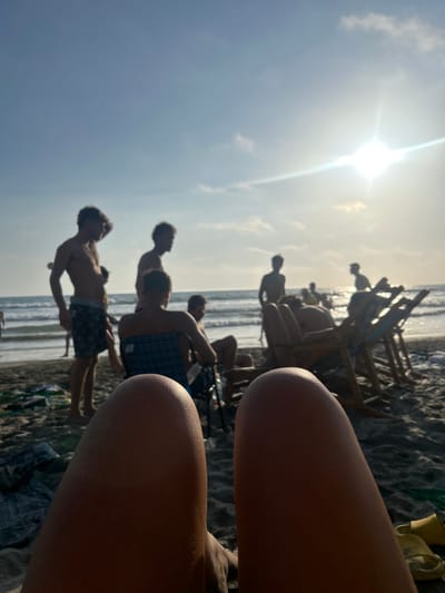 The image shows a close-up view of a person’s legs, seen from the perspective of a person lying on a beach. The legs are tanned and are the main subject of the image. The background is out of focus, and shows a group of people standing and sitting on a beach. Some of the people are wearing swimsuits. The sun is shining brightly in the sky, and the ocean is visible in the distance. The scene appears to be in a beach in Montanita, Ecuador (coordinates -1.825, -80.755), a town known for its surfing and relaxed atmosphere. The environment is a sandy beach, with chairs and other beach accessories scattered around. The people in the background appear to be enjoying themselves and spending time together. The image captures a sense of relaxation and enjoyment.