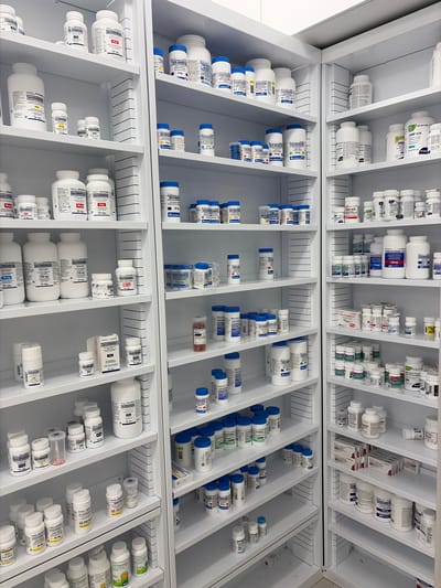 The image shows a retail pharmacy in Longueuil, Canada, specifically inside a store. The pharmacy features a large, white shelving unit filled with various medications in bottles and boxes. The shelves are organized with many white, blue-capped, and clear containers of pharmaceuticals, along with some cardboard packaging. The lighting appears to be standard indoor artificial light. There are no people visible in the image. The overall impression is one of a well-stocked and organized pharmacy interior.