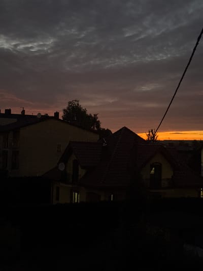 The image captures a twilight scene in Marki, Poland, focusing on the silhouettes of buildings against a dramatic sky. The sky displays a gradient of colors, transitioning from a deep grey with textured clouds at the top to warm oranges and yellows near the horizon. The foreground is dominated by the dark shapes of rooftops, with a prominent multi-gabled roof in the center, suggesting residential architecture. Some buildings show subtle hints of interior light emanating from windows. A thick black cable, possibly a power line, runs diagonally from the upper right corner, adding a stark graphical element to the composition. The overall impression is one of a quiet evening in an urban or suburban environment, with the natural beauty of the sunset juxtaposed against the man-made structures.