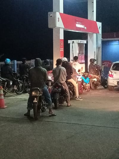 The image depicts a nighttime scene at a gas station in Cimaung, Indonesia. Several individuals, primarily men, are seen waiting in a queue on their motorcycles to refuel. The dark sky indicates the late hour, with the gas station's artificial lighting illuminating the area.

In the foreground and middle ground, a line of motorcyclists faces towards the fuel pumps. They are dressed casually, with some wearing helmets. One visible motorcycle license plate reads "G 3820 ZOB". To the left, behind orange traffic cones and a blue metal fence, more motorcycles are parked. A white car is also visible at the far right, suggesting it is also part of the queue.

The gas station features a white and red structure with pumps. A prominent sign above the pumps displays "PERTAMAX 92" along with the Pertamina logo, identifying the fuel type and company. Below this, a circular red sign clearly states "JALUR KHUSUS," meaning 'Special Lane.' Another partially visible sign to the right appears to direct "PENGENDARA JALUR MOTOR" (Motorcycle Lane Riders). Faint text that seems to read "PER MUR" is also discernible on a small sign or sticker near one of the pumps or motorcycles. The overall atmosphere appears calm, with people patiently waiting for service.