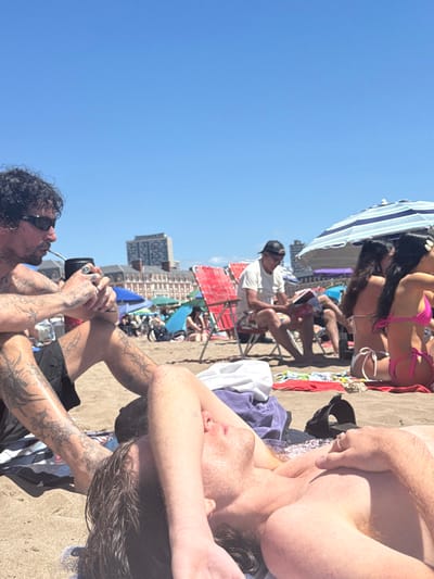 A sunny beach scene with many people. A woman in a pink bikini sits under a striped umbrella. A man in a white shirt and cap sits on a red folding chair. Closer, two men are on the sand, one with tattoos on his arms drinking from a mate gourd, the other fair-skinned with freckles and dark hair. A building is visible in the background.