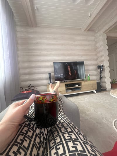 The scene is an interior view within a wooden-walled structure in what appears to be Areyskoye, Russia. A person is holding a dark, floral-patterned mug, likely seated. The individual is wearing patterned pants, resting on a gray surface, probably a couch.

In the background, a modern television displays the image of a woman. A media console holding various devices is beneath the TV. The room has light-colored, presumably wooden, walls, and the ceiling has a decorative trim. A partial view of a window is present in the lower left.