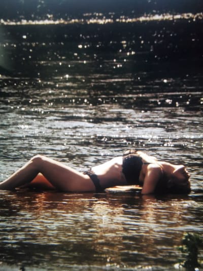 A person is lying on their back in shallow water, possibly a river or stream, with their head resting on the water. They are wearing a black bikini. The sun is reflecting off the water, creating a dappled effect. The background is dark and out of focus, with hints of trees or foliage. The overall impression is one of relaxation and enjoyment of a warm day. The time of day appears to be late afternoon or early evening, given the warm tones of the light on the water and the shadows. The weather is likely pleasant and warm, suitable for swimming.
