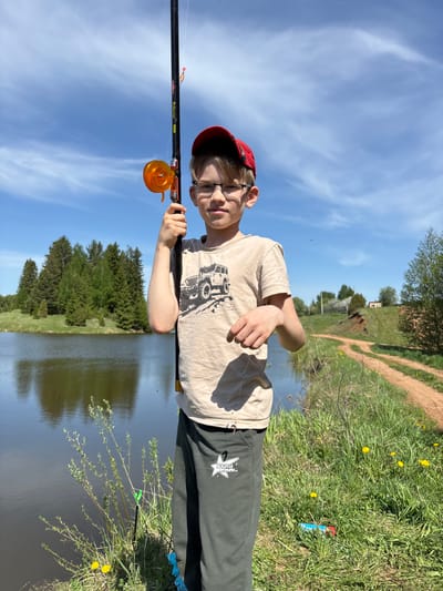 A young boy wearing a red cap and glasses stands at the edge of a lake. He holds a fishing rod with an orange reel. The rod has the name "Princess 500" written on it. The boy is wearing a light-colored t-shirt with a graphic on it and dark-colored pants with a white logo. The lake is surrounded by green grass and some yellow dandelions. A dirt path is visible in the background. The sky is blue with some clouds. The location is most likely Karashur, Russia.