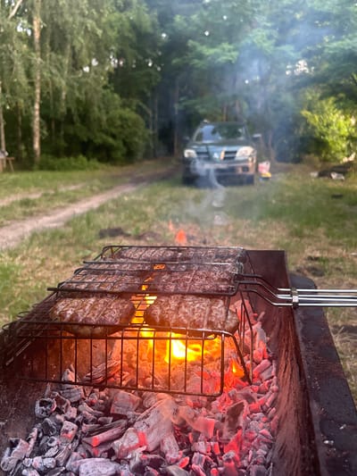 The media file depicts an outdoor barbecue scene. The primary subjects are several rows of meat, likely kebabs or similar preparations, cooking on a metal grill over glowing red hot coals. The grill is positioned within a dark metal barbecue pit, with smoke rising from it and into the air.

In the background, a dirt road winds through a grassy area with scattered trees. Further down the road, a light-colored vehicle, possibly a van or RV, is visible amongst the trees, partially obscured by smoke. The overall setting appears to be a rural or natural environment, likely a campsite or a picnic area.

The activity taking place is the grilling of meat, suggesting a meal preparation or a social gathering centered around food. The time of day seems to be late afternoon or early evening, given the lighting and the presence of smoke, which is more noticeable in lower light conditions. The weather appears to be warm and suitable for outdoor cooking.

Notable details include the intense heat emanating from the coals, evident in the bright orange and yellow flames and the glowing embers. The texture of the meat on the grill is also visible, suggesting it is in the process of being cooked. The presence of the vehicle in the background hints at a possible camping trip or outing. No text is visible in the media file. The overall mood is one of outdoor leisure and culinary activity.
