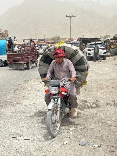 A man wearing a red turban and a purple traditional tunic rides a red Honda motorcycle laden with a large, cylindrical object wrapped in grey tarpaulin. The object appears to be made of stacked tires. He is on a dusty, paved road, likely in the city of Bostan, Pakistan, with other vehicles, including a blue cargo truck, a red three-wheeled cargo truck with "GAPON CARGO" written on it, and a white SUV, visible in the background. Buildings and stalls, suggesting a market area, are present on the sides of the road. Mountains are visible in the distance, shrouded in dust or haze. The overall scene depicts a busy street with commercial activity.