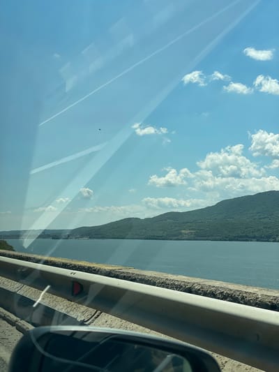 The image depicts a landscape viewed from the interior of a vehicle, positioned alongside a large body of water with forested hills beyond, near Drobeta-Turnu Severin, Romania. The clear blue sky, visible in the upper two-thirds of the frame, is dotted with scattered white cumulus clouds. Two small, dark, irregular specks are present against the sky, one in the upper center and another lower and to the right. The vehicle's windshield exhibits prominent diagonal light reflections and faint geometric reflections, partially obscuring the view. A subtle, thin white line, possibly a contrail reflection, is visible in the upper left.
The middle ground features a wide expanse of dark blue-green water, consistent with the Danube River in the specified location. On the far side of the water, densely forested green hills or mountains rise, extending across the entire background. Faint structures, potentially industrial or related to power generation (consistent with the Iron Gates I Hydroelectric Power Station nearby), are discernible on the distant left bank.
In the foreground, a light gray metallic guardrail runs horizontally across the lower portion of the image, atop a textured, weathered concrete barrier. A red, triangular reflector is affixed to the guardrail, pointing to the left. The reflection of a dark car side mirror is visible in the bottom right corner, revealing a partial view of the road surface and the vehicle's interior. The ground immediately adjacent to the guardrail appears to be rough concrete or asphalt.