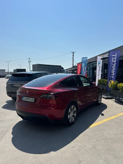 The image displays an outdoor scene in a parking area under a clear blue sky, illuminated by bright sunlight which casts distinct shadows. In the foreground, a red Tesla Model Y is parked with its rear facing the viewer, displaying a Bulgarian license plate "BG EA 8899 AB". Behind the Tesla, a dark grey Range Rover is parked, also showing a Bulgarian license plate "BG CB 6726 TH". In the background, several large semi-trailer trucks are visible, indicating a commercial or transport-oriented environment. To the right, a building features several tall, vertical advertising flags. These flags prominently display the logos and names of major Turkish telecommunication companies: a red flag with "Vodafone" (partially visible), a white flag with "Türk Telekom", and a blue flag with "TURKCELL". The combination of Bulgarian-registered vehicles and prominent Turkish brand advertising strongly suggests a location in Edirne Merkez, Turkey, likely a service station, shopping area, or border facility frequented by international travelers. The ground consists of paved asphalt or concrete, and utility poles with power lines are visible against the horizon. No people are clearly identifiable in the scene.