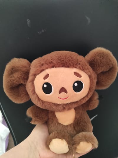 A plush toy resembling a brown monkey with large, round ears and a lighter brown face is held in a person's hand. The toy has big, black eyes with white pupils, small brown eyebrows, and a red smiling mouth. Its body is covered in soft, brown fur, and it has light brown paws. The toy is held from the side, with its head facing upwards and its body extending downwards and to the left. The background is a dark, flat surface, possibly a table. A person's hand, with light skin and a subtle ring on one finger, is visible holding the toy, suggesting it is being presented or played with. The image is taken from a slightly high angle. No specific time of day, weather, or further location cues are apparent beyond the provided context of Constanța, Romania. No visible text is present in the image.