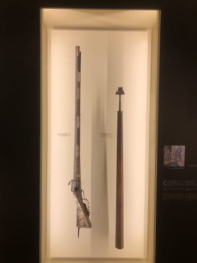 This image depicts two firearms displayed in a museum exhibit, likely located in Veytaux, Switzerland. The exhibit features a long, wooden-barreled weapon with an arrow-like head and a more ornate firearm with an intricate metal design along its barrel. The exhibit includes accompanying text, possibly providing information about the weapons' history and usage. The firearms are showcased inside a glass case with a bright, neutral background, contrasting with the dark surroundings of the museum. There's also a smaller illustration visible at the top of the display.