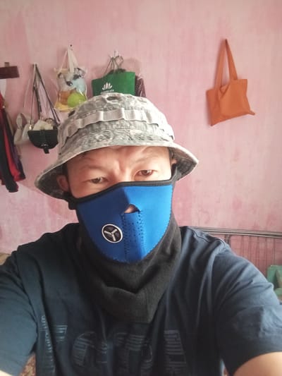 A close-up, medium shot captures a person from the chest up, facing forward and looking directly at the camera. They are wearing a camouflage bucket hat and a bright blue face mask that covers their nose and mouth, with a black fabric extending down their neck and chest. The mask features a circular emblem with a white design. The person is wearing a dark blue t-shirt with a faint, dark gray graphic.
The background consists of a pink wall with some items hanging on it. To the left, there are several bags and what appear to be articles of clothing hanging on hooks. One bag is clear and contains stuffed toys and round objects. Further to the left, a dark object, possibly a bag or purse, is visible. In the center background, a green reusable shopping bag with a white lotus flower logo and text is hanging. To the right, an orange tote bag is also hanging on the wall. The wall has a textured, slightly uneven surface. To the far right, a metal rack or barrier is partially visible.
The scene appears to be indoors, likely within a residential setting. There are no clear indicators of a specific location in Kojomkul, Kyrgyzstan, other than the possibility that the person or items might be associated with that region. The overall lighting is somewhat dim, suggesting an interior light source.