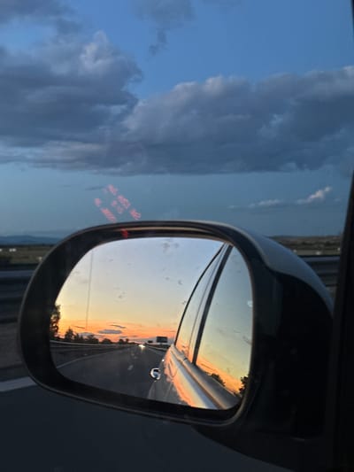 The image captures a view from a vehicle's side mirror, reflecting a sunset over a highway in Manolsko Konare, Bulgaria. The sky is painted with hues of orange, pink, and yellow, contrasting with the dark, dramatic clouds overhead. The highway stretches into the distance, with white lines marking the lanes and a few vehicles visible. Trees line the side of the road, their silhouettes sharp against the vibrant sky. The reflection in the mirror is clear, showing a portion of the car's interior and the passenger-side window. Text, seemingly from a display on the car, is visible as red digits and letters in the upper part of the reflection, showing "OUT AUTO AG2" and "R:15" and "SO:38888". The overall scene evokes a sense of travel and the beauty of a sunset.