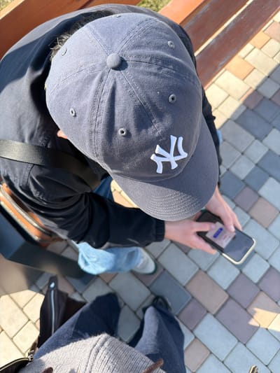 An overhead, high-angle shot captures two individuals outdoors in Verin Ptghni, Armenia. The primary subject, seen from the upper back, wears a dark grey baseball cap with a white "NY" logo, a dark jacket, and light blue jeans. This individual is seated on a wooden bench, with their back partially visible, and is actively engaging with a smartphone held in both hands, its screen illuminated. A black strap from a bag or garment crosses their left shoulder. The secondary subject, who appears to be the photographer, is partially visible in the foreground, showing their lower torso, clad in a light grey knitted top and dark pants, with a dark bag positioned near their feet. The ground surface is paved with rectangular interlocking pavers in varying shades of grey, beige, and light brown. Strong directional sunlight casts distinct shadows across the scene.