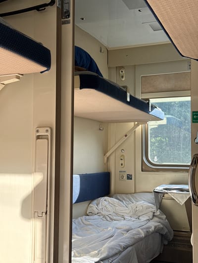 The image depicts the interior of a sleeper cabin on a train. The scene is brightly lit by daylight filtering through a window. The cabin contains two berths: an upper berth partially visible at the top of the frame and a lower berth that is configured as a seating area. A blue cushion rests on a small section of the floor beside the lower berth. A white comforter is rumpled on the right side of the lower berth. The opposite side of the lower berth has a hinged seat back in the upright position. The cabin wall features electrical outlets, a window with a partially obscured view to the outside, and a ventilation vent. The location in the city of Kirpichnoye, Russia, is not evident from the image.
