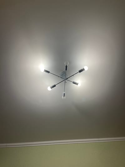 An interior photograph captures a ceiling-mounted light fixture with five exposed bulbs, arranged in a starburst pattern. The bulbs are illuminated, casting a bright light and creating a gradient effect on the white ceiling. Below the ceiling, a decorative white cornice molding separates it from a light green wall with a subtle textured pattern. The scene appears to be in an indoor residential or commercial setting. The image does not provide any information that would allow for its location to be identified as Muravlenko, Russia.