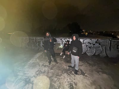 The image captures three individuals at night on an elevated concrete surface in the Gerakas Municipal Unit, Greece. A male with a beard stands on the left, wearing a dark hooded jacket with a grey upper portion and dark pants, presenting a thumbs-up gesture with his right hand. A second male stands in the right foreground, wearing a black hooded sweatshirt with the hood up, light grey sweatpants, and a black Adidas shoulder bag. A third individual, partially obscured behind the second male and seated on a low concrete structure, is looking down at a small illuminated device. Behind the subjects is a concrete wall heavily covered in dark-colored graffiti. Beyond the wall, a distant city landscape with scattered lights is visible under a dark sky. The ground is a textured, discolored concrete surface. Circular lens flare artifacts are prominent in the upper left and lower central areas of the image.