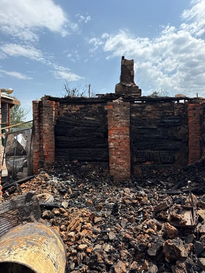 The image depicts the remains of a structure in Nedryhailiv, Ukraine, severely damaged by fire under a bright, partly cloudy sky. The central subject is a partially standing brick-walled building, approximately three to four meters in height, with its interior completely gutted. The bricks, predominantly reddish-brown, show significant charring and soot staining on their inner surfaces, particularly where wooden elements once existed. Horizontally arranged charred wood or logs are visible within the wall sections, indicating former structural components. A damaged, upright brick chimney structure remains at the top of the rear wall, slightly off-center to the right.

The ground directly in front of and within the destroyed structure is covered by a substantial accumulation of debris, consisting of fragmented bricks, charred wood, ash, and other unrecognizable burnt materials. In the lower-left foreground, a large, cylindrical metal object, possibly a barrel or tank, lies on its side, exhibiting rust and burn marks. Near it, a section of black mesh material is visible. In the lower-right foreground, amidst the rubble, a small, rectangular, severely charred object with metallic components is present.

To the left of the main damaged structure, a portion of an adjacent building with a corrugated metal roof and an overhead light fixture is visible, appearing less affected by fire damage. A distorted, reflective, and crumpled sheet material, possibly insulation, is propped near this adjacent structure. The background includes sparse, leafless vegetation and a slender, vertical pole-like structure. No people, active actions, or interactions are present in the scene.