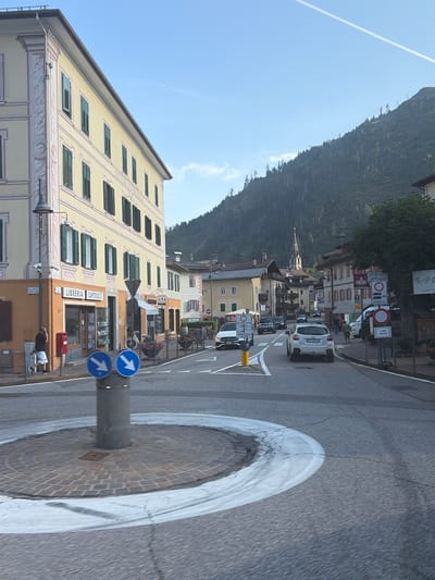 The image captures a street scene in Predazzo, Italy, likely on a clear day with a bright blue sky and a faint contrail. In the foreground, a circular roundabout is visible, paved with stone tiles and outlined by white markings on the asphalt road. A gray cylindrical pole stands in the center of the roundabout, topped with two circular blue signs, each displaying a white downward-pointing arrow.
To the left of the roundabout, a multi-story building with a pale yellow facade and decorative pink trim dominates the view. The building features numerous windows with dark green shutters. At the base of this building, a shop with a sign reading "LIBRERIA CAPITOLO" is visible, with books displayed in its window. A red mailbox is positioned next to the shop entrance. A man in a dark shirt and shorts stands near the building, appearing to be on his phone.
The street itself is paved with asphalt and shows signs of wear, with white lane markings and some visible tire marks. Several cars are present on the road, including a white SUV in the middle lane and other vehicles further down the street. Buildings line the opposite side of the street, featuring a mix of architectural styles and colors, with some exhibiting pitched roofs. A church spire with a cross is visible in the distance, rising above the rooftops.
The background of the image is dominated by a steep, tree-covered mountainside. The trees are dark green, creating a dense forest that stretches up the slope. The overall scene depicts a typical European town street, with a blend of residential and commercial buildings set against a natural mountain backdrop.