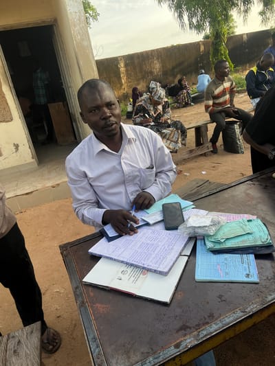 This image depicts an outdoor administrative scene in Gbaza, Nigeria. In the foreground, a dark-skinned man with short dark hair, wearing a light-colored collared shirt, is seated or standing behind a worn, dark metal table. He holds a blue pen in his right hand and looks directly towards the viewer. His left hand rests on an open, spiral-bound notebook filled with blue handwritten text. A black smartphone lies face down on the table to his right, next to a clear plastic bag containing small items. Further right on the table are stacks of various documents, including light blue and green booklets, and a blue form labeled "TRANSPORT CORPORATION OF NIGERIA."

To the left of the main subject, a partial view of a person wearing black pants and brown sandals is visible. In the background, behind the main subject, is an open doorway of a light-colored, worn building, revealing a person in a plaid shirt inside. Further in the background, against a tall, light-colored concrete or mud wall, several individuals are gathered. A woman in a patterned dress and headscarf is seated, actively writing in a notebook. To her right, a man in a striped polo shirt (brown, orange, white) is seated on a wooden bench, holding a dark object. Other individuals are present, including a person in a light blue tunic seated on a bench further back, and a man in a blue track jacket partially visible on the far right. A tree with green foliage is visible above the background wall. The ground is sandy and earthy.