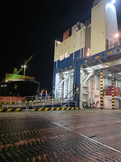 At night in the city of Ravenna, Italy, a ferry is docked at a port. The foreground shows a wet, textured deck with chevron patterns, likely made of metal, with some red and black streaks indicating wear or fluid. Beyond the deck, a ramp or gangway with yellow and black striped safety barriers leads towards a large ferry. This ferry, partially visible on the left, is black and has the name "GULLUK" written on its hull. It appears to be a cargo or vehicle ferry. The main structure of the ferry, on the right, is white and blue, with various levels and mechanical components visible. It is illuminated by bright lights, creating strong shadows. Shipping containers, some with visible logos like "UN", are stacked on the upper decks of the ferry. In the background, other docked vessels and port structures are dimly visible, also lit by lights. The overall scene suggests a busy port at night, with the ferry preparing for or having just completed a journey.