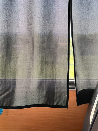 The image shows a close-up of a window covered by two sheer, gray curtains with black trim. A sliver of green foliage is visible through the partially open window. To the right of the curtains is a light brown, wood-paneled wall. The angle of the shot suggests the viewer is positioned slightly below the window. There are no people visible. The location in the city of Бизяки, Russia is not visually identifiable from the image.