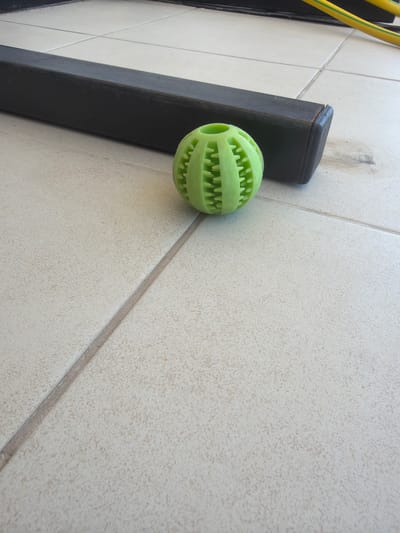 The image shows a light green, textured rubber ball on a tiled floor. A dark rectangular object, possibly a piece of furniture or equipment, is positioned behind the ball. In the background, a yellow and green striped hose is visible. The tiled floor appears to be outdoors, with grout lines visible between the tiles. The overall setting suggests a patio or outdoor area. This image was taken in Sderot, Israel.