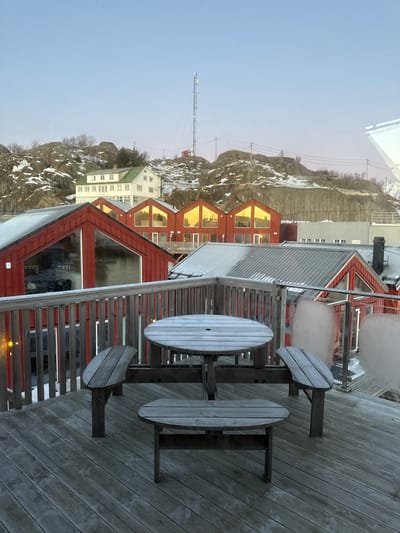 The image depicts a winter scene from an elevated wooden deck in Stamsund, Norway, likely during low light conditions such as dawn or dusk.
In the immediate foreground, a weathered wooden deck extends across the bottom of the frame. A unique, light grey wooden picnic table occupies the center, featuring a circular tabletop with a central circular hole, and three integrated, curved bench seats. One smaller, oval-shaped wooden bench is positioned below the tabletop, closer to the deck surface. To the right, a section of railing features glass panels with translucent, irregular patches of frost or ice adhering to the inner surface. A wooden railing runs along the left side.
Beyond the deck, the midground showcases a cluster of buildings. Directly below the deck railing on the left is a red-painted building with a large, angled window. Further back, a row of three identical red-painted structures with white trim and grey roofs is visible. Each of these buildings presents a large, gabled window reflecting a strong golden light, suggesting direct, low-angle sunlight. The roofs of these buildings are partially covered with snow. To the far right, a larger grey building with a pitched roof is visible.
The background consists of rugged, rocky hills, partially covered in snow and sparse, bare vegetation. A multi-story white building with a distinctive green roof is situated on a lower part of these hills. Dominating the highest point of the terrain is a tall, slender metal communication tower or mast, with a small red structure at its base. Overhead power lines or cables are visible stretching across the hillside. The sky is a clear, pale blue, transitioning to a very faint pinkish hue near the horizon. No individuals are present in the scene.