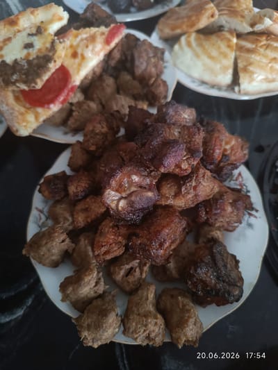 A close-up, overhead shot showcases a feast laid out on a black, reflective surface, likely a table. In the foreground, a white plate overflows with an assortment of grilled meats and small, irregularly shaped baked goods. The meats appear to be a mix of darker, well-browned pieces and lighter, more uniformly cooked morsels. To the left of the plate, a slice of pizza with visible cheese, meat, and tomato is partially obscuring other food items. In the background, another plate holds several pieces of golden-brown bread, sliced and stacked. The lighting is dim, casting subtle shadows and highlighting the textures of the food. A timestamp in the bottom right corner reads "20.06.2026 17:54." The location is identified as Қырғауылды, Kazakhstan.