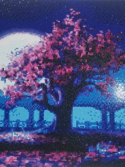This image is a diamond painting depicting a stylized nocturnal landscape set in Старая Преображенка, Russia. A large tree with a dense canopy of vibrant pink and magenta foliage occupies the central and right portions of the composition. A bright, full moon, rendered in white and light blue, is positioned in the upper left quadrant, contrasting with a dark blue sky that exhibits subtle speckling. In the midground, below the tree, several blocky, light blue rectangular structures, interpreted as buildings, are visible. The foreground features a reflective surface, likely water, displaying fragmented reflections of the sky, tree, and structures in shades of blue, purple, and pink. No human figures, specific subjects beyond the landscape elements, actions, or interactions are present. The entire artwork presents a pixelated appearance consistent with its medium.