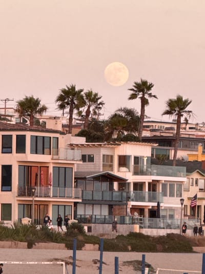 The image captures a twilight scene in Manhattan Beach, California, with a full moon prominently displayed in the pale pink sky. A cluster of multi-story beach houses, characterized by their modern architecture, glass balconies, and large windows, dominates the mid-ground. Several palm trees are scattered among the buildings, their fronds silhouetted against the sky.
In the foreground, a sandy beach area is visible, with a volleyball net and posts suggesting recreational activity. A few individuals are seen walking or standing on the elevated walkways and patios of the houses, as well as on the beach itself. One group of people is gathered on a raised area in front of one of the houses, while others are more scattered. An American flag flies from a pole on one of the buildings. The overall impression is one of a serene evening in a popular coastal community.