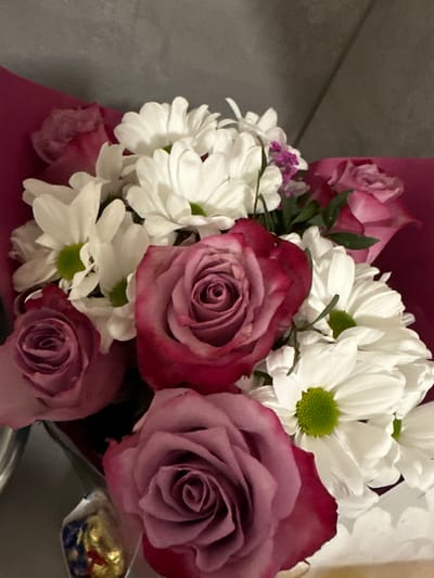 A close-up shot displays a bouquet of flowers. The bouquet contains a mix of roses with a reddish-purple hue and white daisies with green centers. The flowers are arranged in a cluster, with the roses predominating the arrangement, with some small purple flowers interspersed among them. The bouquet is wrapped in pink paper. There is foil wrapped candy in the lower right corner. There are no people visible in the image. The bouquet is possibly in a room with grey walls. The location is unspecified and not obviously Gunzenhausen, Germany.