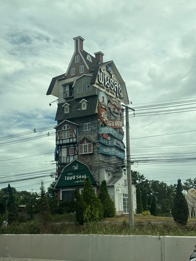 This image captures a unique, multi-story building that resembles a stack of houses, located in the city of Ban Khao Yai, Thailand. The structure, which appears to be a commercial building, features various architectural styles, with gabled roofs, dormer windows, and different facade materials like brick and wood paneling. Prominently displayed on the side of the building are large signs with Thai script, one of which reads "1000 Sook." The building is situated alongside a road with numerous utility wires and poles crisscrossing in front of it. Lush greenery, including trees and bushes, surrounds the base of the building and extends into the background. The sky is overcast with clouds, indicating a dull or rainy day.