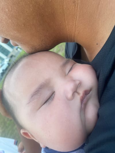 The image shows a close-up of a sleeping baby nestled against an adult's shoulder. The baby's face is mostly visible, with eyes closed and lips slightly parted. The adult's skin, with visible pores and some faint lines, occupies the left side of the frame. A black garment, likely a shirt, is draped over the adult's shoulder and partially covers the baby. The background is blurred, with hints of greenery and what appears to be a railing or fence, suggesting an outdoor setting. The lighting appears natural, implying it could be daytime. The overall mood is peaceful and intimate, capturing a moment of rest for the baby. No text is visible in the image.