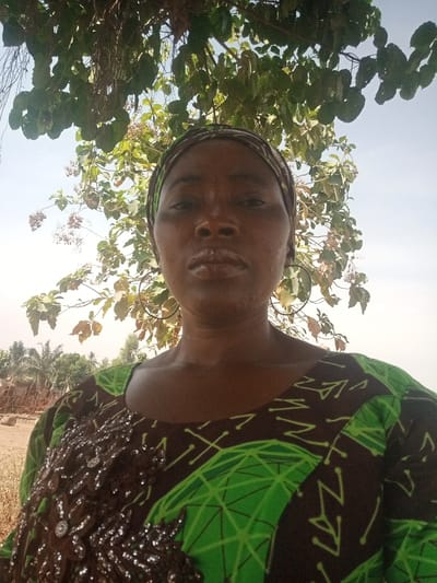 An adult female is depicted in a medium close-up, outdoor, selfie-style photograph in Gudige Sabo, Nigeria. She has dark skin and a neutral expression, looking directly at the camera. Her head is covered by a patterned headscarf, primarily green and dark brown. She wears large, dark-colored circular hoop earrings. Her attire consists of a dark-toned garment featuring bright green geometric patterns and an elaborate, dark, sequined or beaded appliqué on the upper left chest area. Above and slightly behind the subject is a dense canopy of green tree leaves, interspersed with light-colored clusters, against a hazy, light blue sky. The distant lower left background reveals blurred vegetation and rudimentary reddish-brown ground structures.