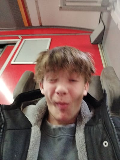 A young person is seated on what appears to be a train. The individual is wearing a gray sweater beneath a dark jacket with a light-colored fur lining. The person has brown hair and is making a comical, puckered face. The backdrop reveals a red interior with framed panels and overhead fixtures, typical of a train carriage. The headrest of the train seat can also be seen.
