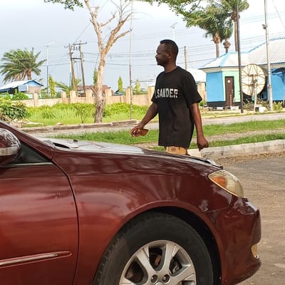 This outdoor scene in Koko, Nigeria, features an adult male positioned next to a reddish-brown sedan. The male, with short dark hair and dark skin, is wearing a black t-shirt with "JSANDER" printed in white text and light-colored shorts. He holds a brown smartphone-like object in his left hand and is looking towards the right side of the frame. The sedan's front-right quarter is visible, including a silver multi-spoke wheel, an amber headlight, and a portion of the hood, which has a partially obscured, light-colored object resting on it. The vehicle is situated on a paved surface adjacent to a curb and a verdant median strip. The background reveals abundant green foliage, several palm trees, a prominent bare-branched tree, and utility poles with overhead lines. In the distance, two light-blue buildings with white roofs are visible, one featuring a large metallic satellite dish-like structure on a pole. The sky appears overcast.
