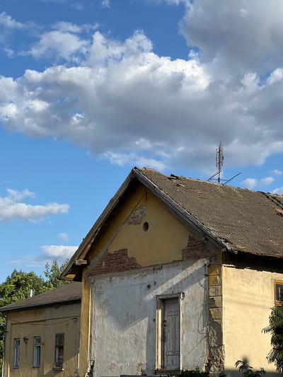 The image shows the exterior of a dilapidated building, possibly a house, in Újezd u Brna, Czechia. The building has a weathered, yellow facade with sections of exposed brick and crumbling plaster. A rustic, light-colored wooden door is visible on the front of the structure. The roof is covered with corrugated material and appears to be in disrepair, with some tiles missing. A tall antenna, likely for telecommunications, is mounted on the roof. The sky above is blue with scattered white clouds. Lush green trees can be seen in the background. The overall impression is one of neglect and age. There are no people visible in the image.